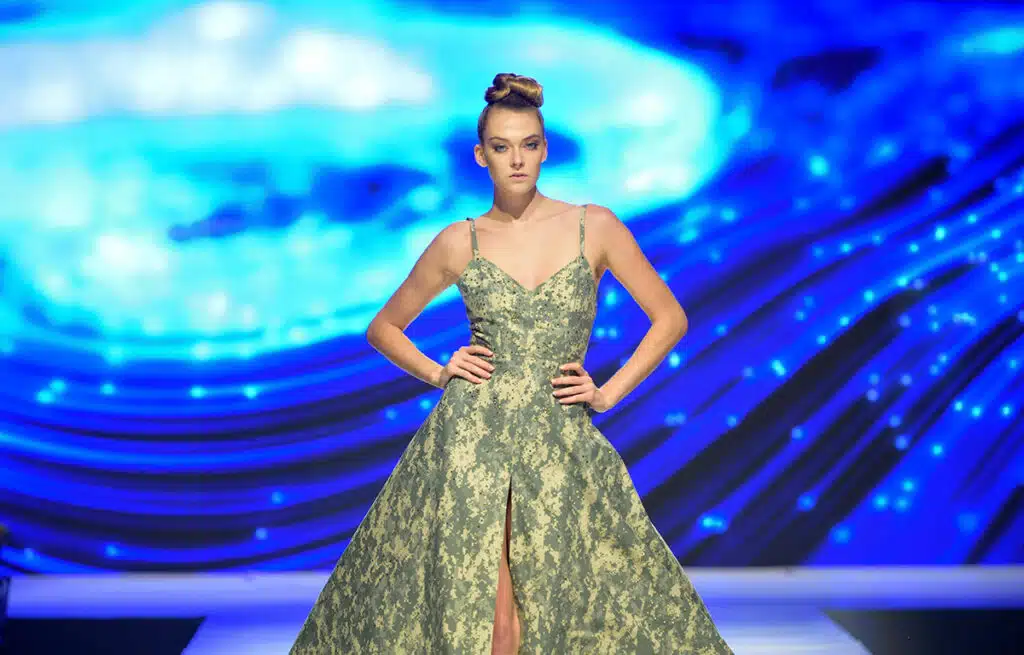 A model in a green floral gown with a thigh-high slit walks confidently on a runway. The background features vibrant blue, swirling lights, creating a dynamic atmosphere.