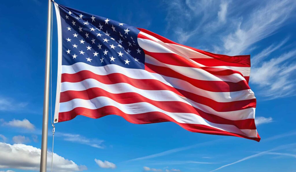 The image shows the flag of the United States waving against a blue sky with some clouds. The flag features red and white stripes and a blue field with white stars.