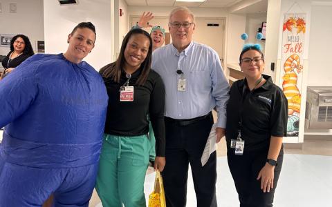 A group of four people stands together in a hallway. One person is wearing a blue inflatable suit, and another is dressed in medical scrubs. They are smiling, with a "Hello Fall" banner in the background. A fifth person in a blue wig waves from behind.