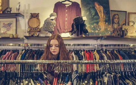 A person with long red hair looks through clothing racks in a vintage store. The shop is decorated with framed paintings, art pieces, and a sweater hanging on the wall above. Various colorful garments are displayed on hangers.