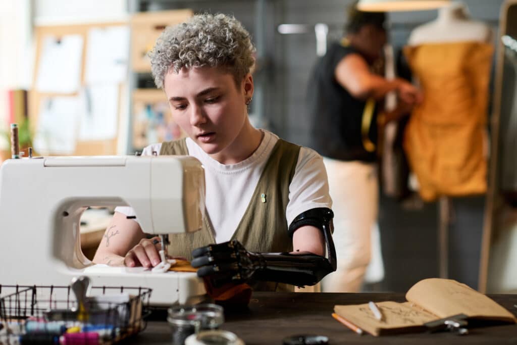 A person with short curly hair and a prosthetic arm concentrates on sewing fabric with a sewing machine. In the blurred background, another person is adjusting a dress on a mannequin. A sketchbook and sewing materials are on the table.